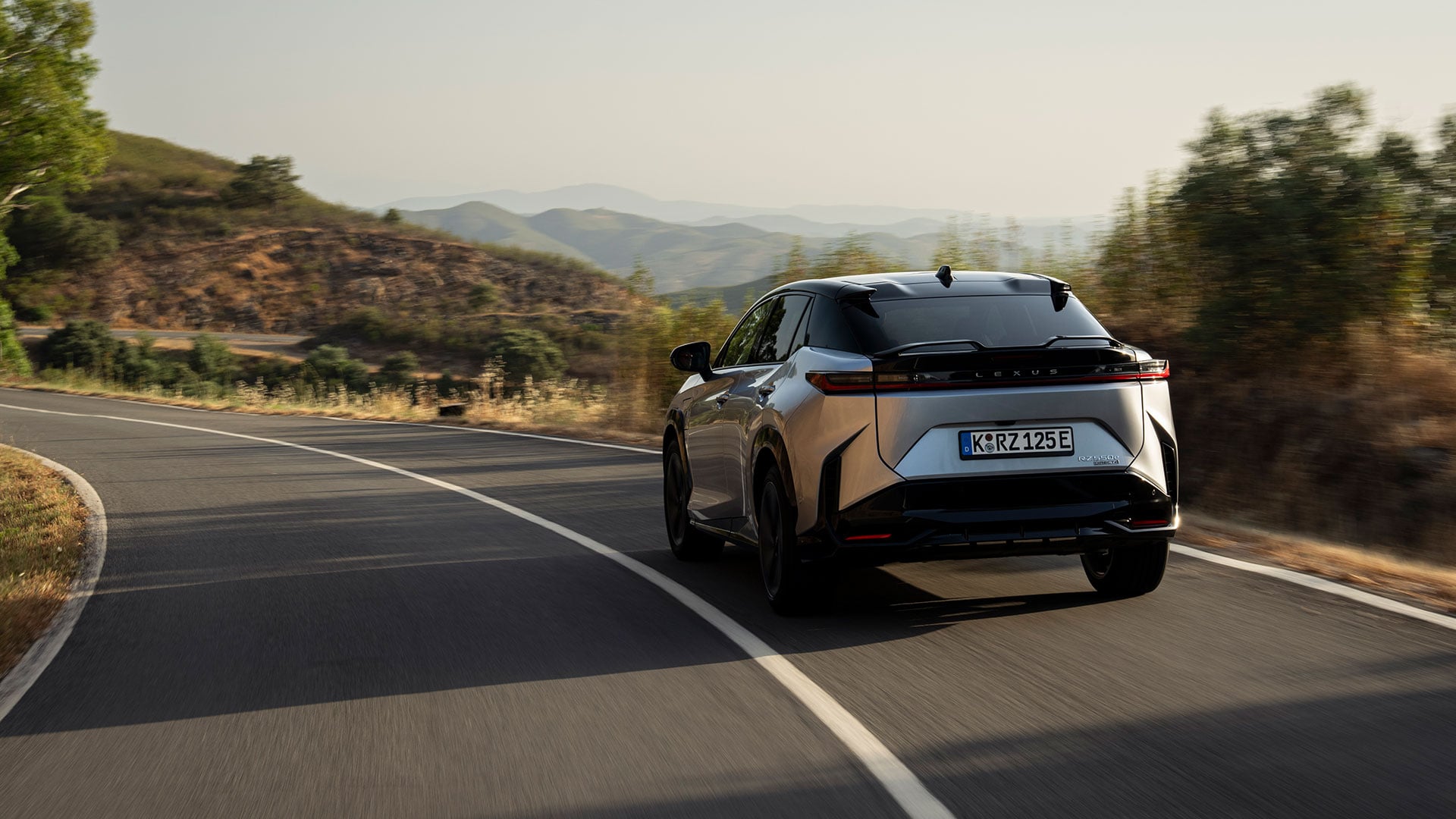 Silver Lexus RZ electric SUV driving on a winding road through a natural landscape
