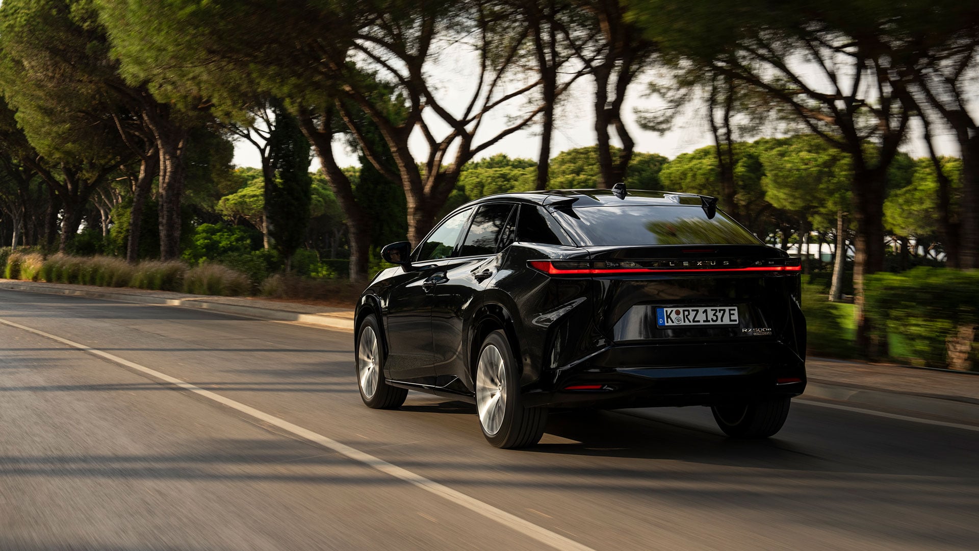 Rear view of a black Lexus RZ electric SUV driving along a tree-lined road