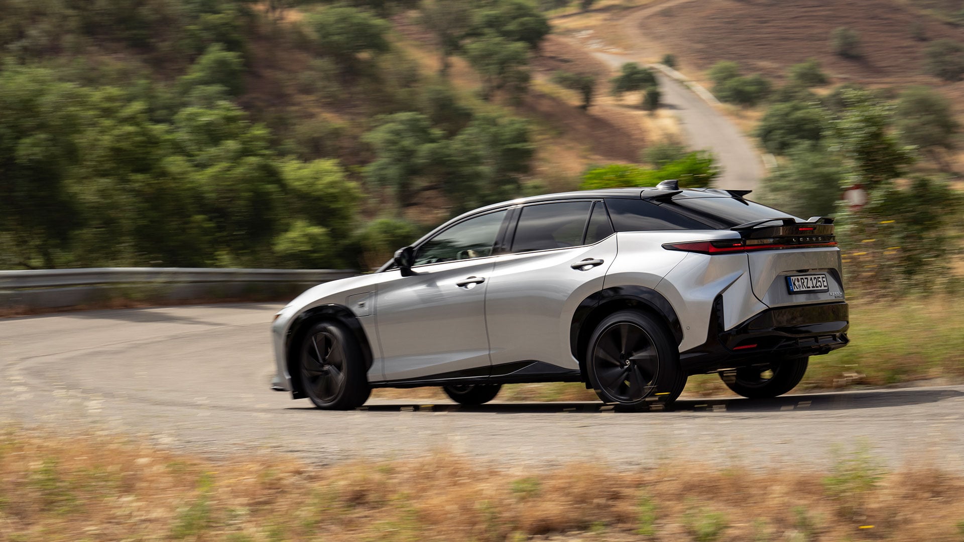 Silver Lexus RZ electric SUV driving along a winding road through a rural landscape